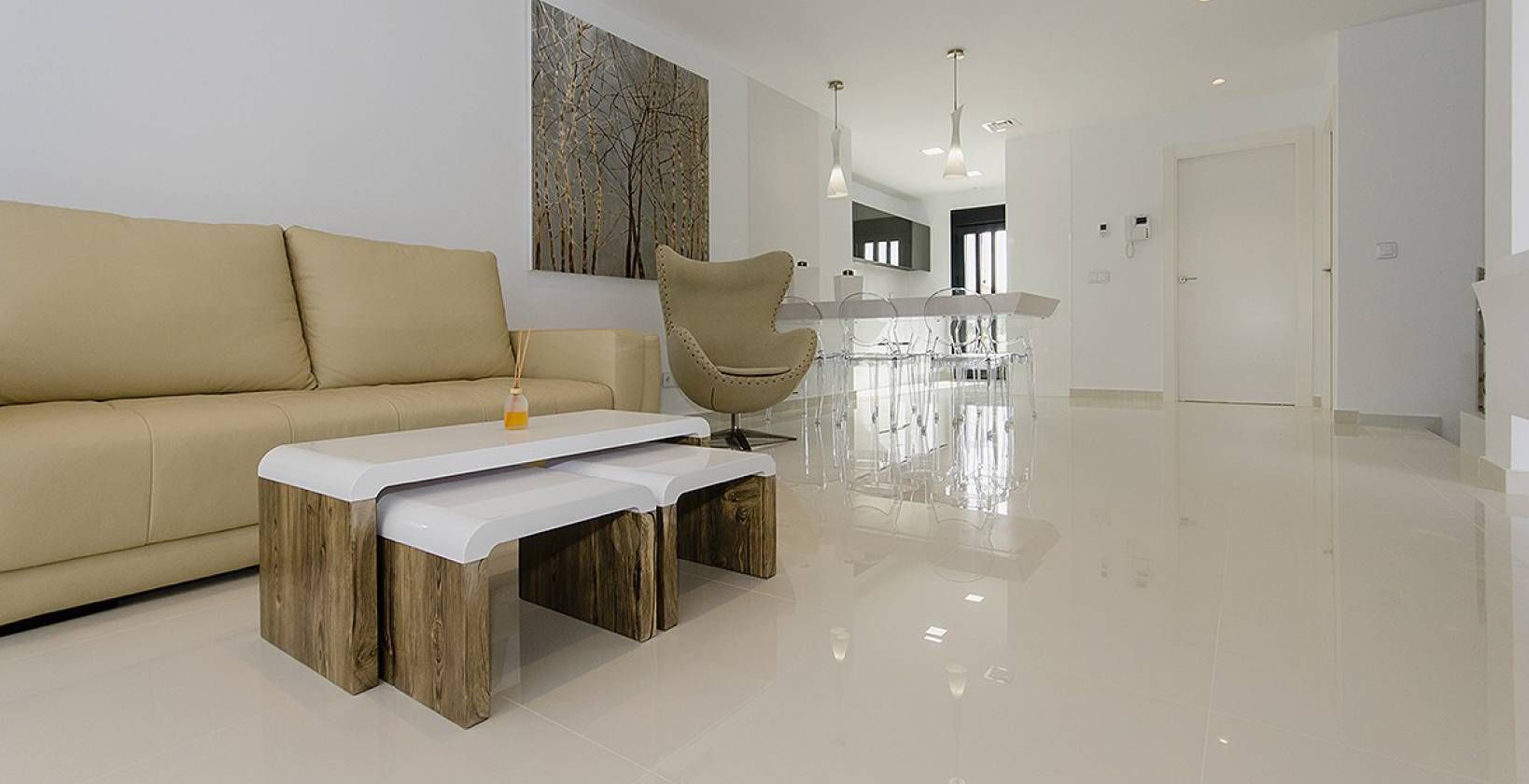 Open-plan living area with a beige sofa, nested wooden-and-white coffee tables, and a clear dining set under pendant lights.