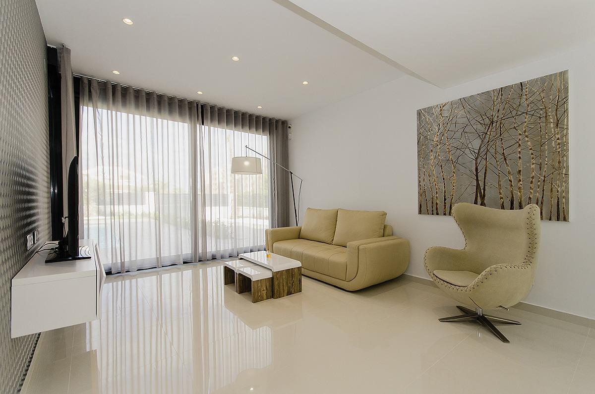 Modern living room with a beige sofa, curved armchair, glass coffee table, and birch-tree artwork on a white wall by large windows with sheer curtains.