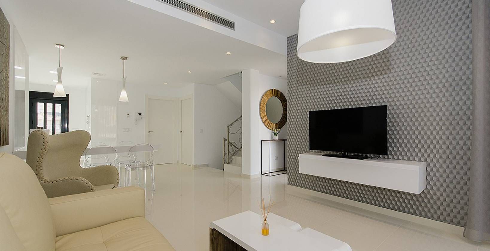 Open-plan living room with a glossy white floor, beige armchairs, transparent dining chairs, and a textured gray wall behind a wall-mounted TV.