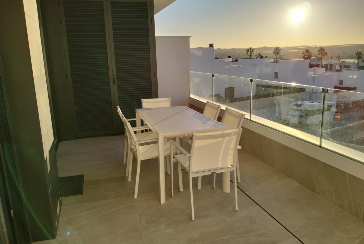 White outdoor dining set on a modern balcony with glass railing, overlooking white buildings and palm trees against a low sun.