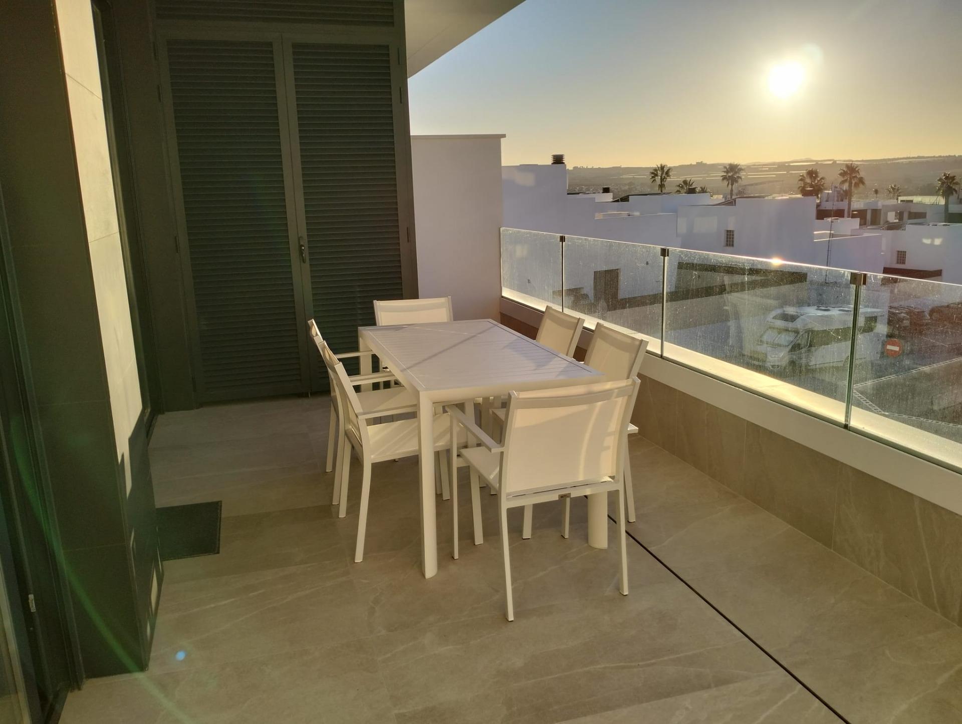 White outdoor dining set on a modern balcony with glass railing, overlooking white buildings and palm trees against a low sun.
