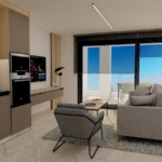 Open-plan living area with a beige kitchen, gray seating, and a balcony with an ocean view through large glass doors.