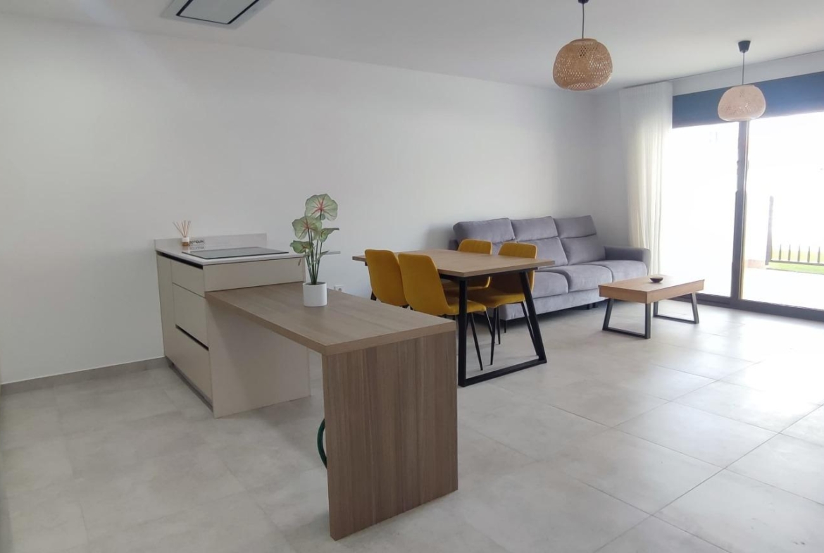 Open-plan living room with gray sofa, wooden dining table with yellow chairs, a small potted plant, and a large glass door filling the space with light.