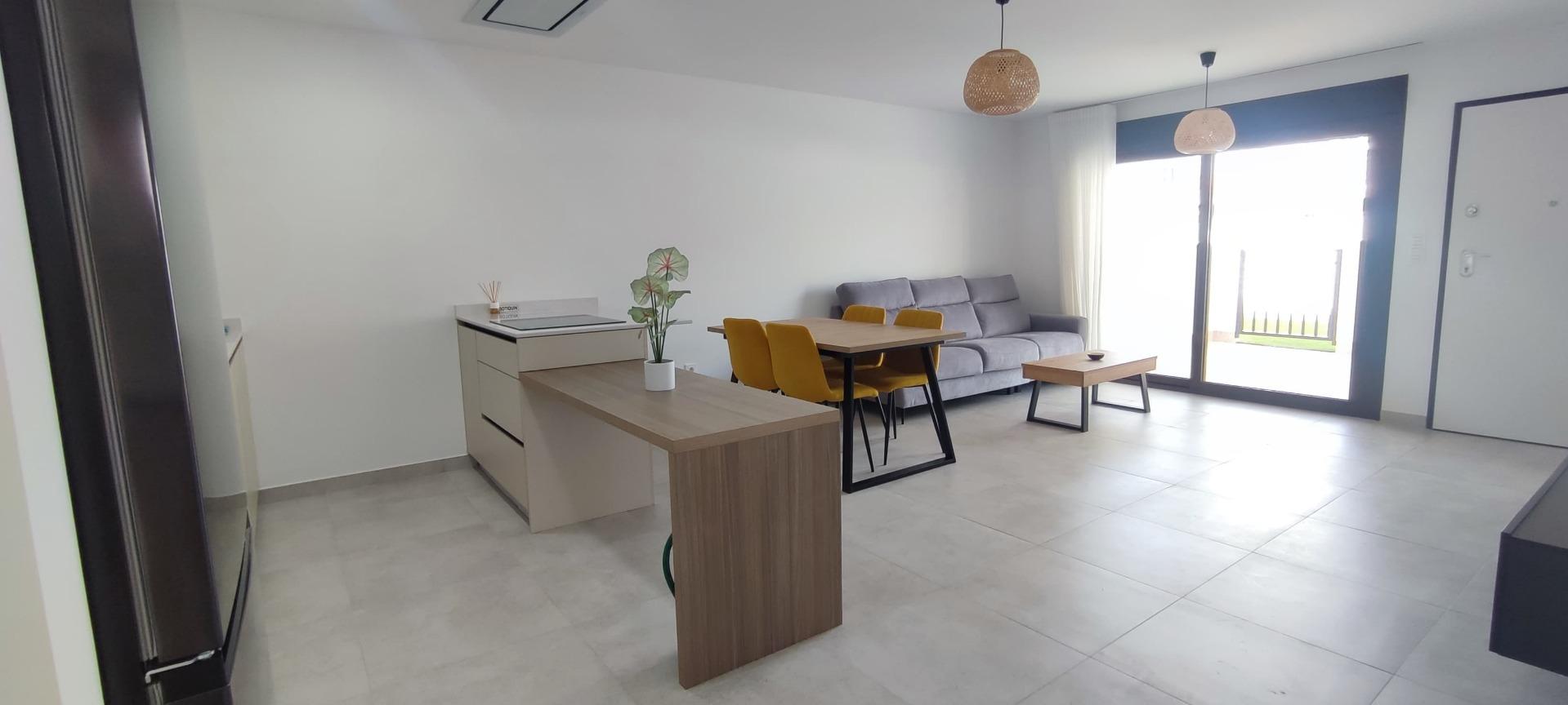 Open-plan living room with gray sofa, wooden dining table with yellow chairs, a small potted plant, and a large glass door filling the space with light.