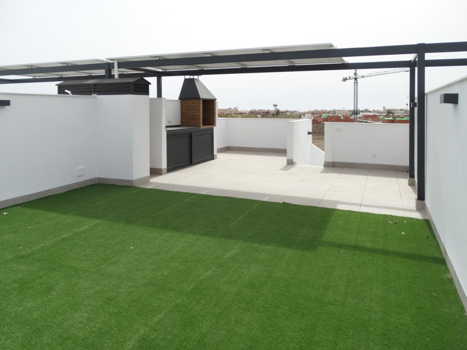 Rooftop terrace with green artificial grass, white walls, and a brick barbecue under a metal canopy, with city buildings visible in the distance.