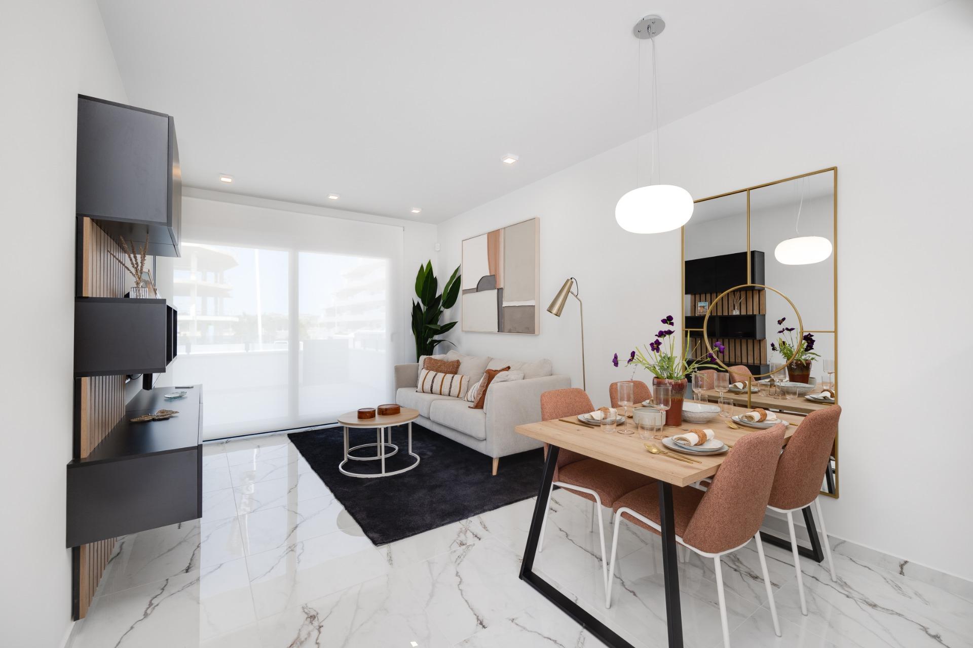 Open-plan living and dining area with a white marble floor, gray sofa, black rug, and a wooden dining table with pink chairs and a gold-framed mirror on the wall.