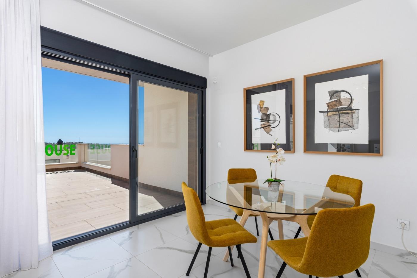 Bright dining area with a round glass table, four mustard chairs, white marble floor, and two abstract framed prints; sliding glass doors open to a sunlit balcony with blue sky.