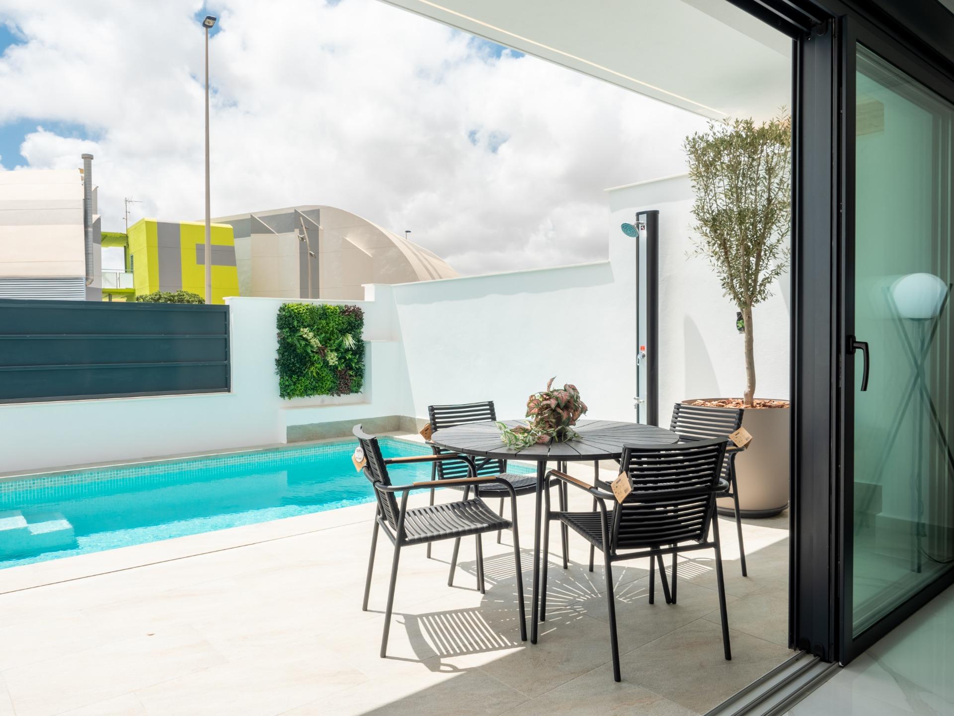 Poolside patio with a black metal dining table and chairs beside a turquoise pool, glass doors on the right, and modern buildings in the background