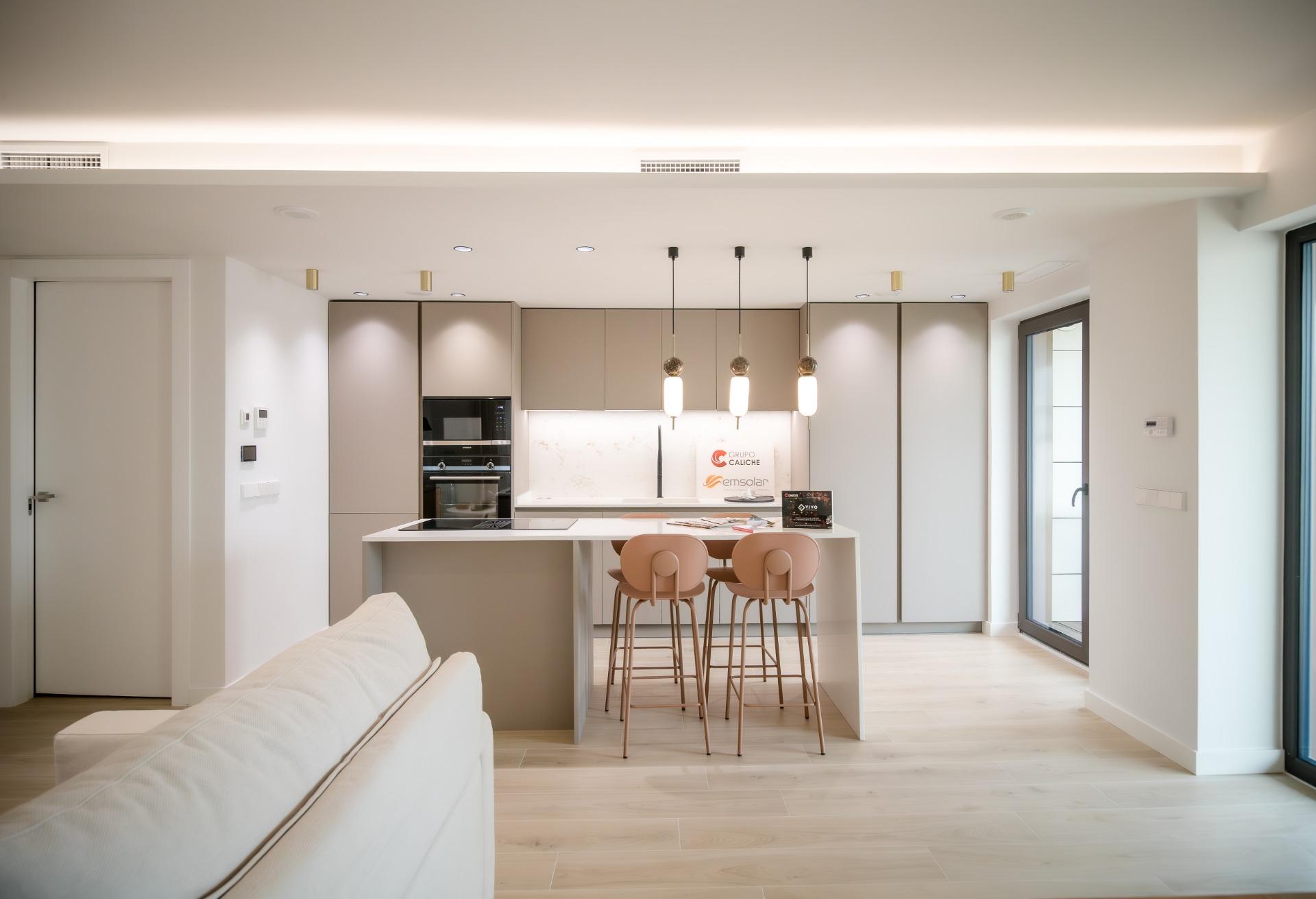 Open-plan kitchen with beige cabinets, marble backsplash, and a white island with pink-beige bar stools under pendant lights.