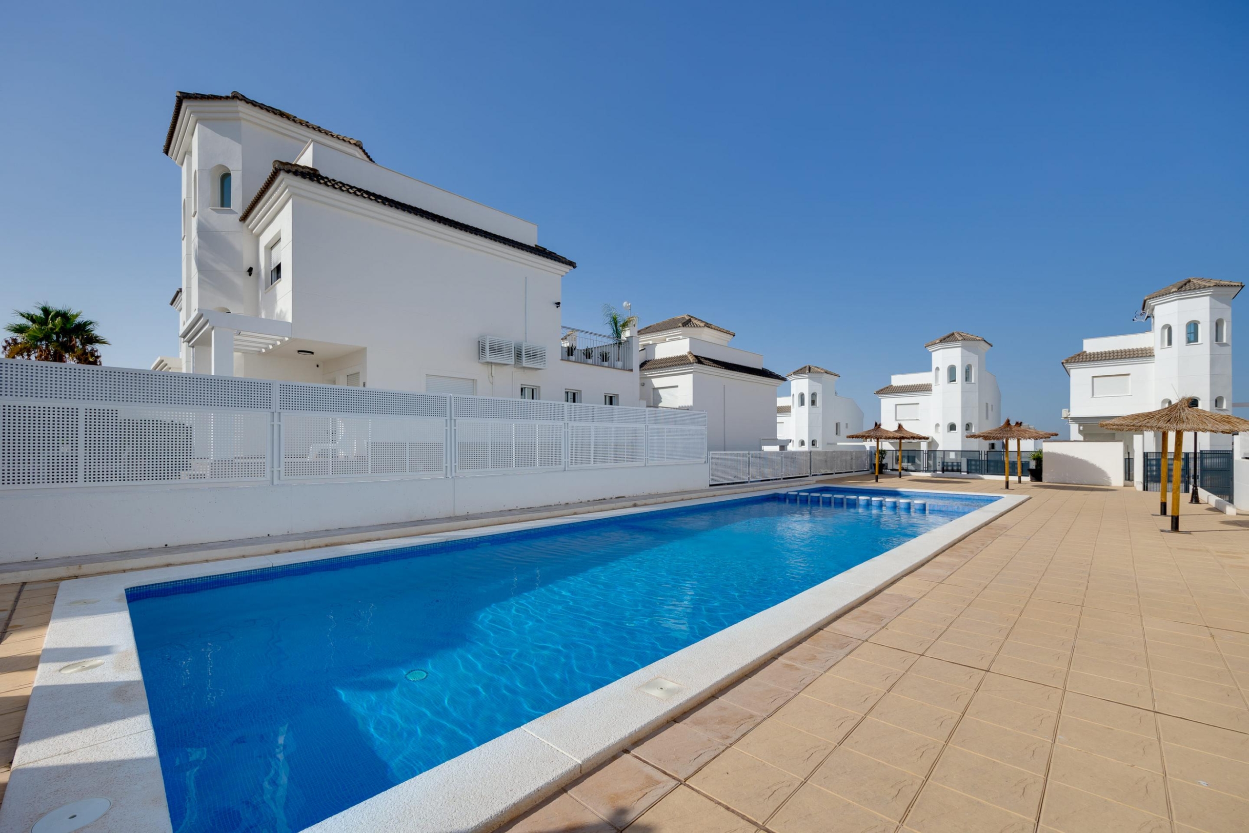 White Mediterranean-style buildings surround a long blue swimming pool under a clear sky, with straw umbrellas along the deck.