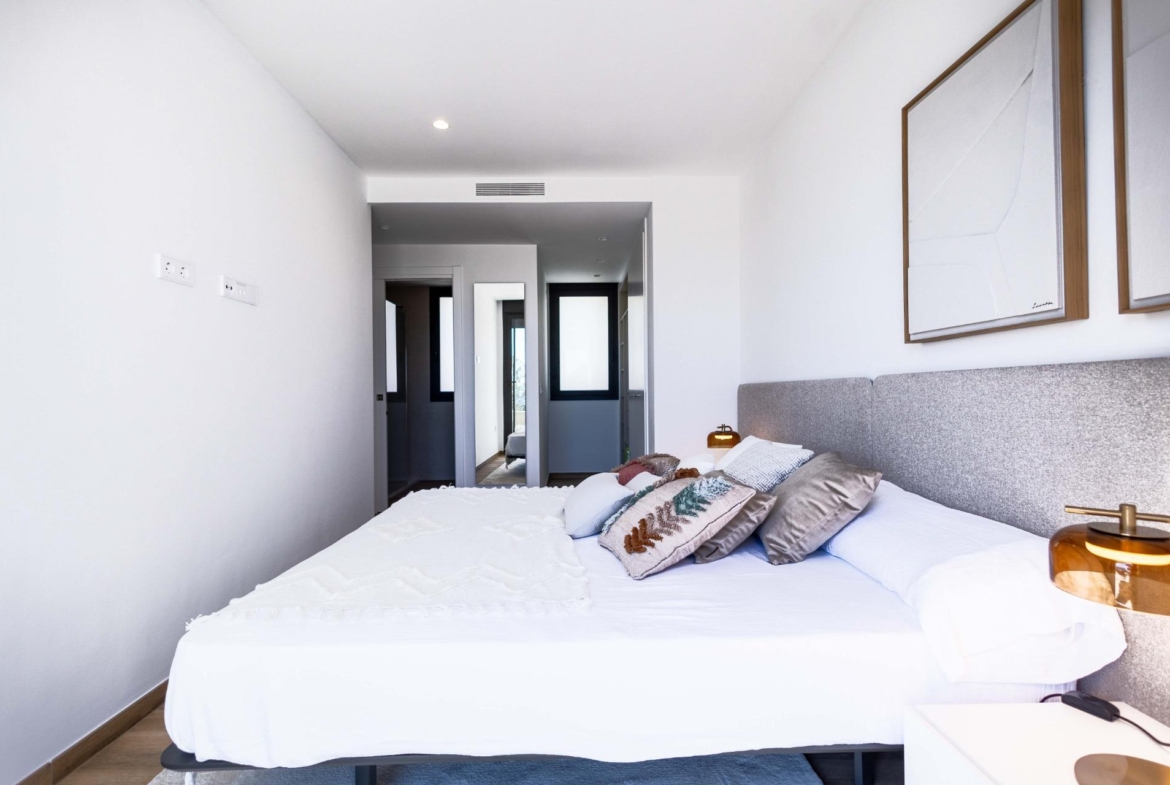 A modern white bedroom with a large bed, pillows, and a gray headboard; a hallway visible in the background through an open doorway.