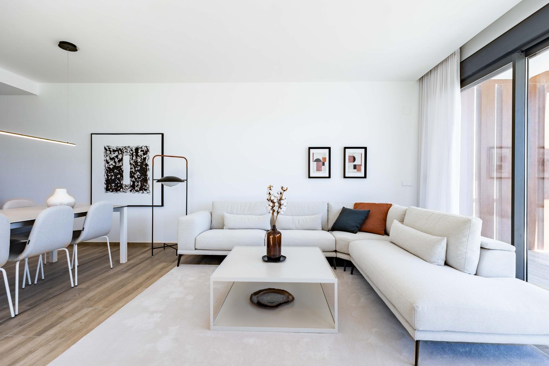 Bright, modern living room with a pale sectional sofa, white square coffee table, and large glass doors with sheer curtains.