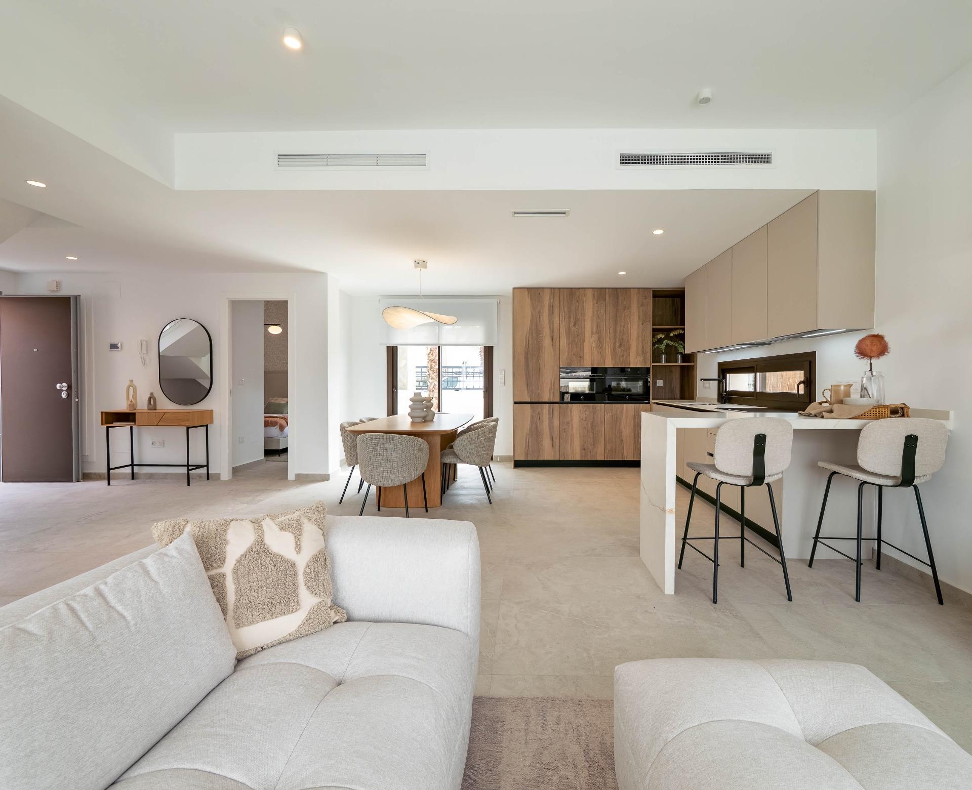 Open-plan living area with a light-gray sofa in the foreground, dining table and upholstered chairs, and a wooden-kitchen with built-in cabinets in the background.