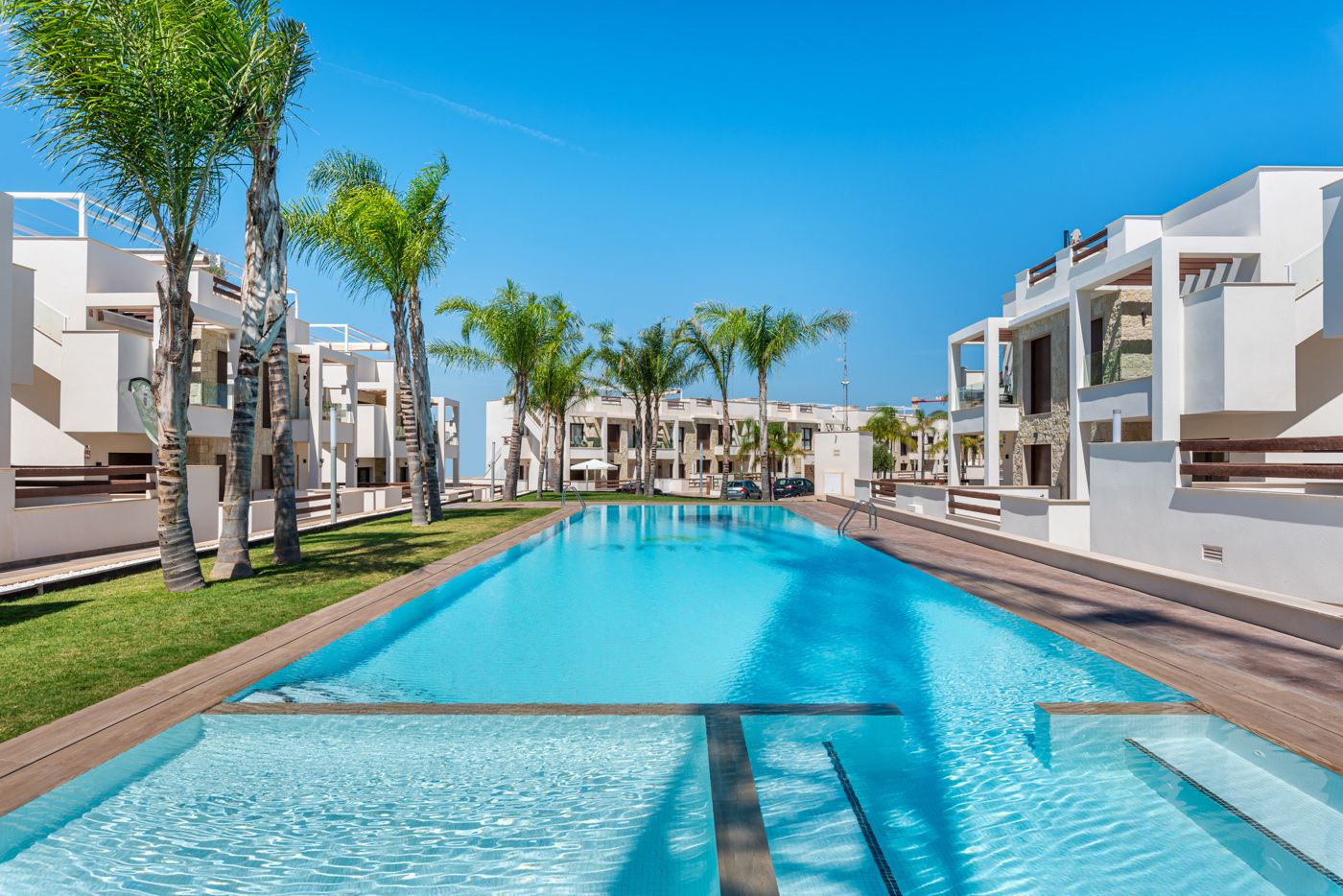 Resort-style residential complex with a long rectangular pool, white buildings, and palm trees under a clear blue sky.