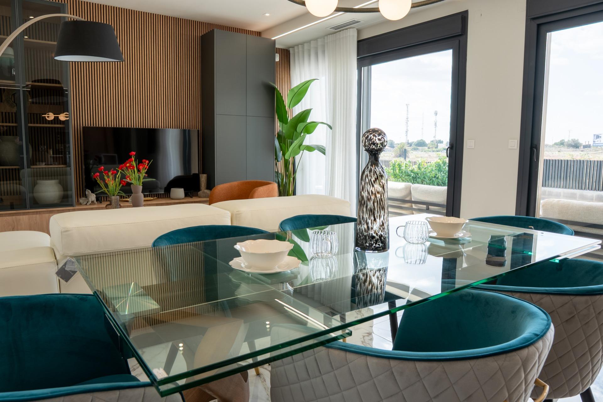 Open-plan dining area with a glass table, white sectional, teal chairs, and large windows offering an outdoor view.