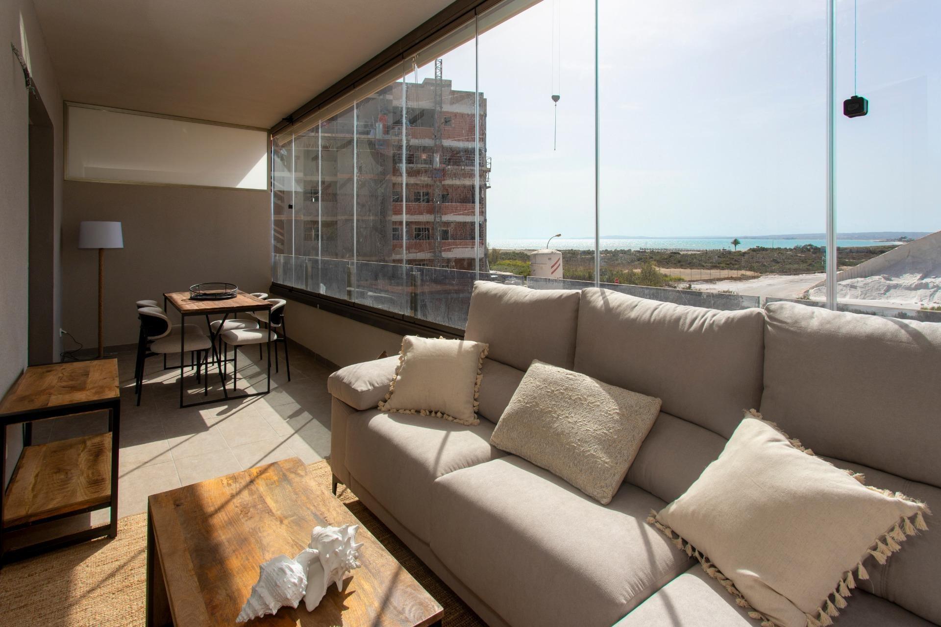 Sunlit living room with a beige sectional sofa and wooden coffee tables facing a long glass wall overlooking the sea.