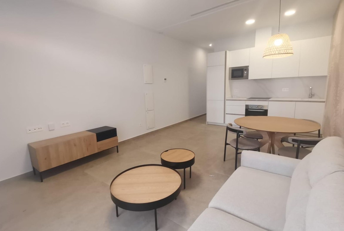 Open-plan living area with a white kitchenette, round wooden dining table, and light gray sofa. Two round nesting coffee tables sit on a concrete floor against a minimalist backdrop.