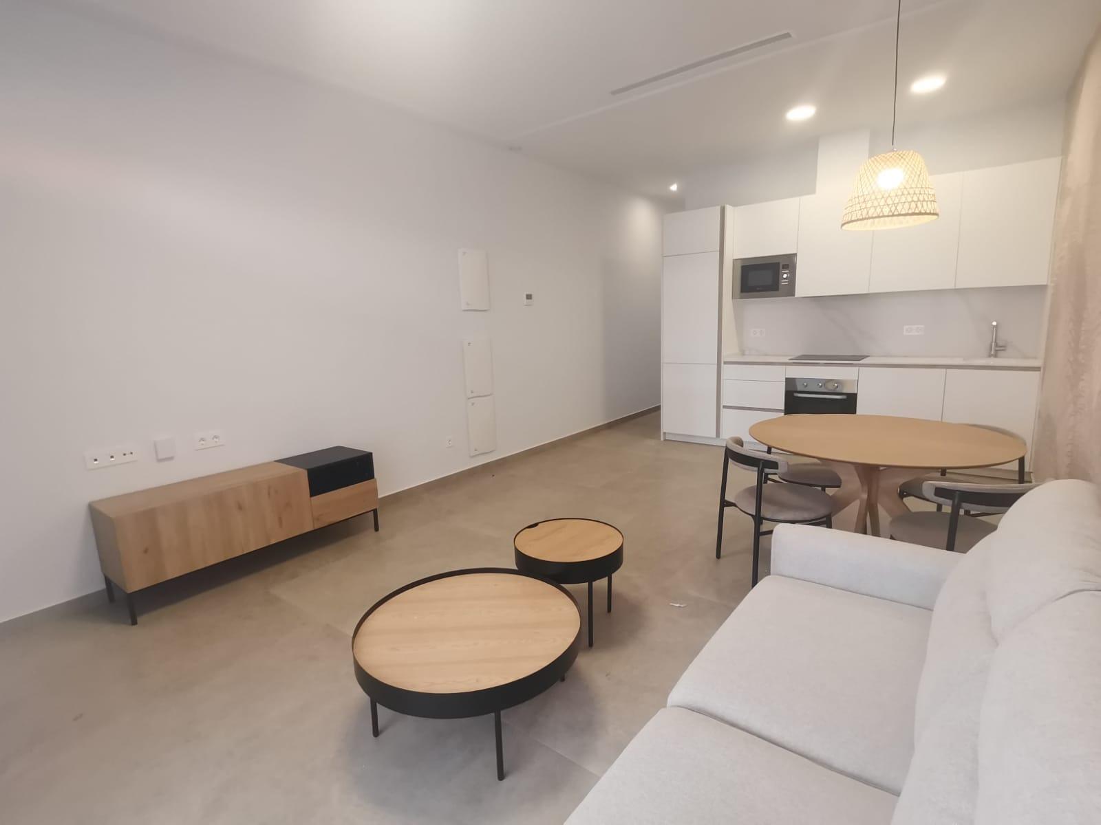 Open-plan living area with a white kitchenette, round wooden dining table, and light gray sofa. Two round nesting coffee tables sit on a concrete floor against a minimalist backdrop.