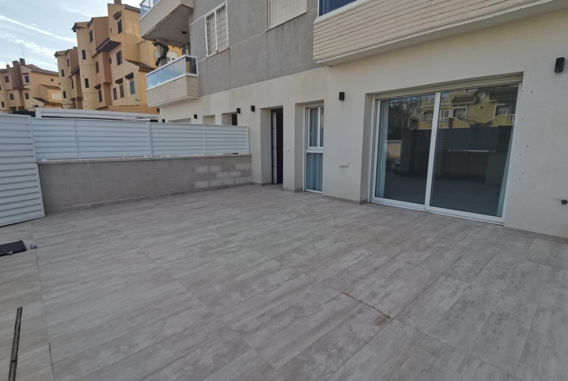 Ground-level terrace of a modern apartment building with sliding glass doors, white walls, and a gray tiled floor.