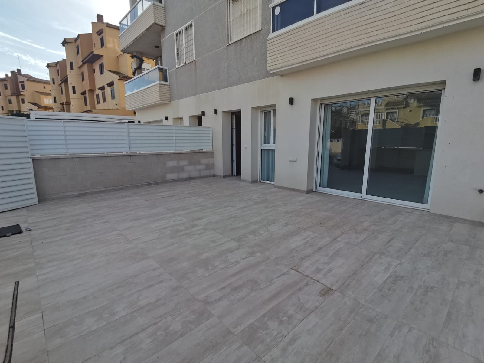 Ground-level terrace of a modern apartment building with sliding glass doors, white walls, and a gray tiled floor.