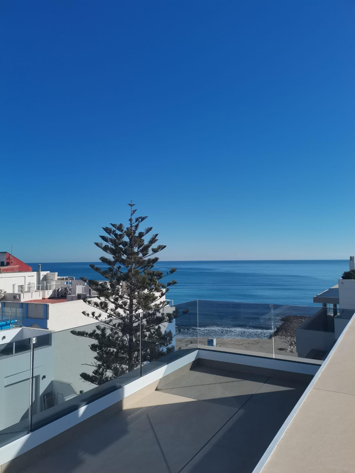 View from a balcony: glass railing, modern white buildings, a tall evergreen tree, and a calm blue ocean under a clear sky.