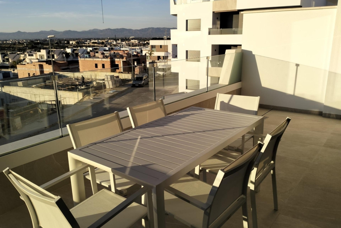 Rooftop balcony with a long white dining table and chairs, glass railing, overlooking a city and distant mountains at sunset.