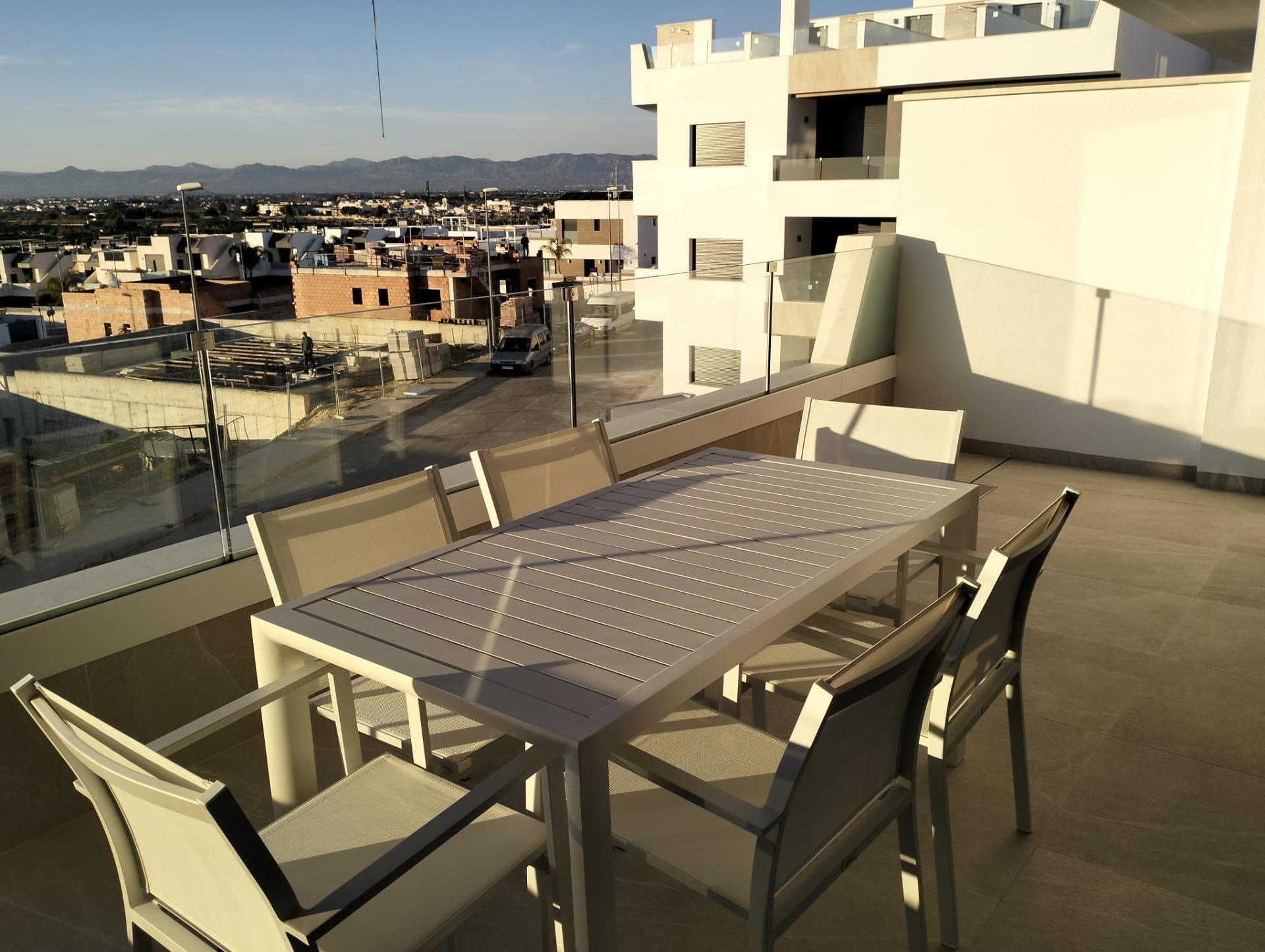 Rooftop balcony with a long white dining table and chairs, glass railing, overlooking a city and distant mountains at sunset.