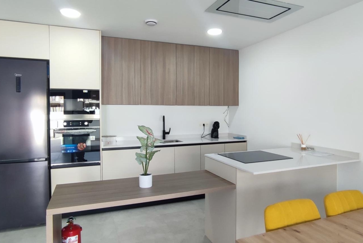 Modern kitchen with tall black refrigerator, built-in oven, beige lower cabinets, and wood-finish upper cabinets along a white wall.
