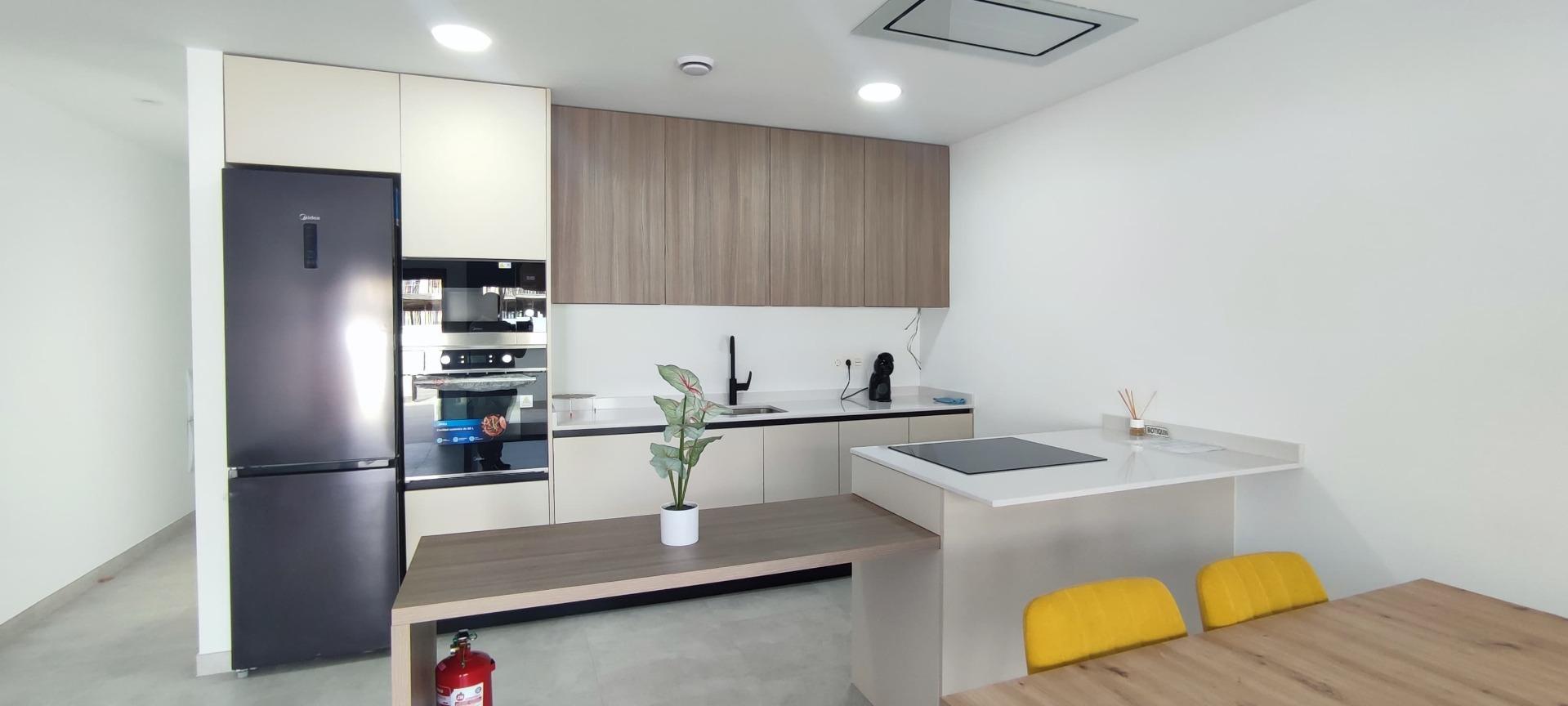 Modern kitchen with tall black refrigerator, built-in oven, beige lower cabinets, and wood-finish upper cabinets along a white wall.