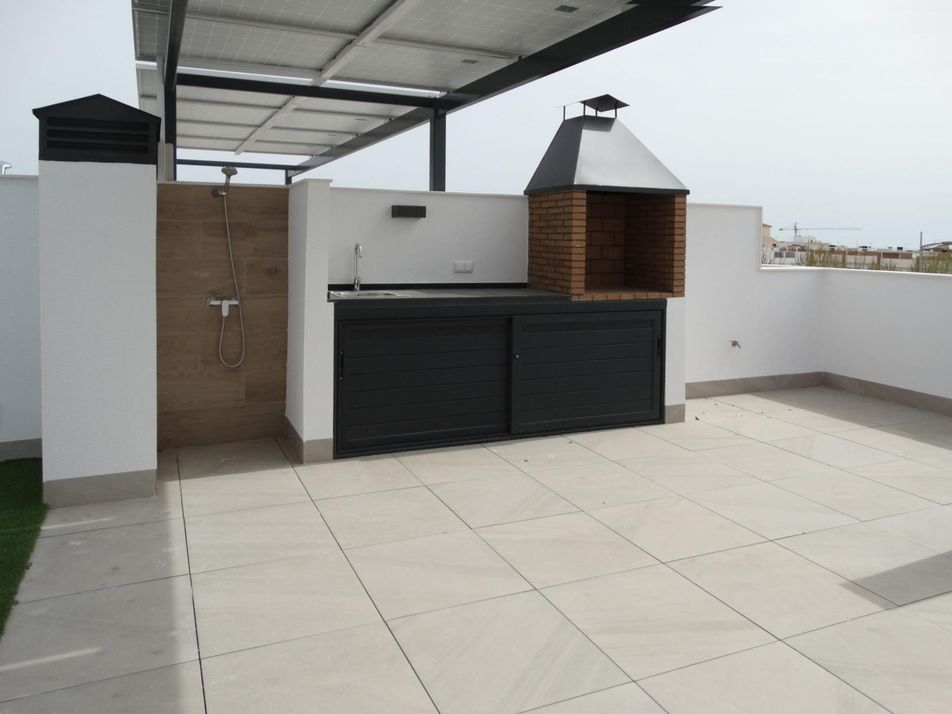 Rooftop outdoor kitchen with brick barbecue under a metal hood, sink and dark cabinet on a tiled terrace.