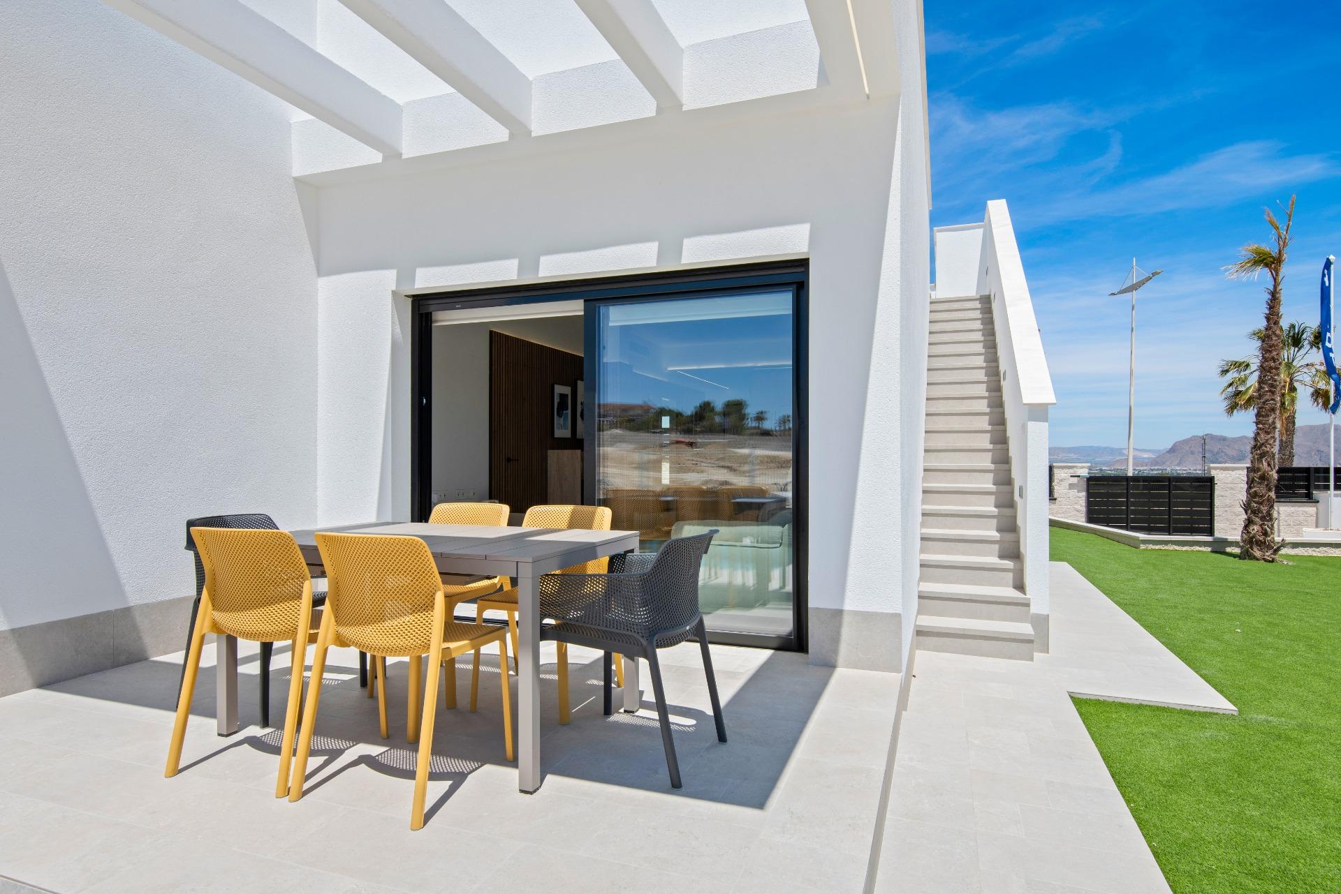 Modern white patio with a gray dining table and yellow and black chairs under a pergola, sliding glass door to indoors, and stairs on the right.