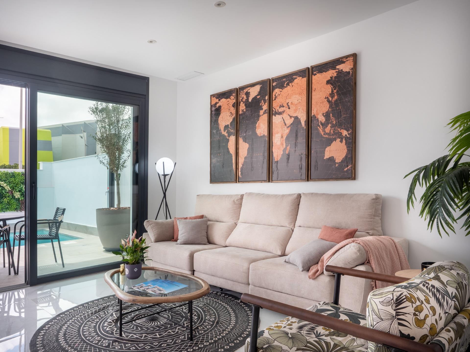 Living room with a beige sectional sofa beneath a four-panel copper world map wall art, a glass coffee table, and a black geometric rug. A sliding glass door to a patio is on the left. There are potted plants nearby.