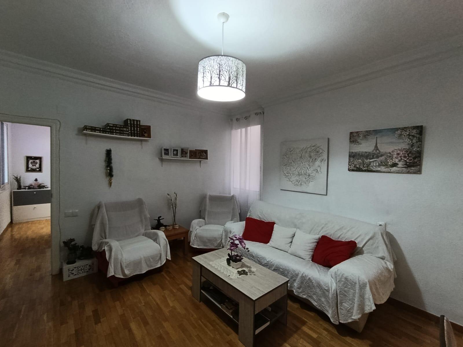 Cozy living room with white sofa and chairs, red cushions, wooden floor, wall art, and a tree-pattern ceiling lamp.