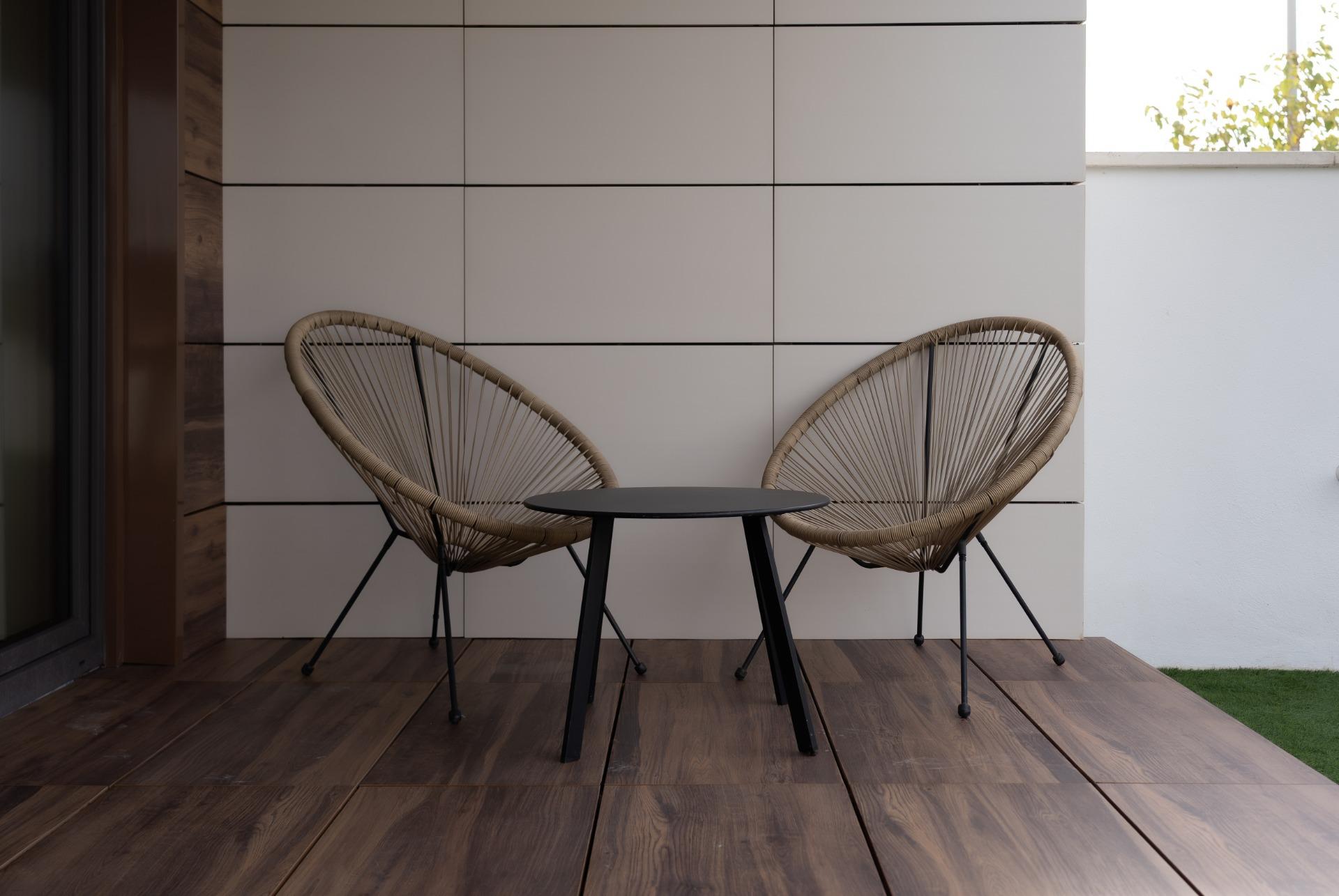 Two woven rattan chairs flank a small black round table on a wooden patio with a white tiled wall behind it outdoor seating.