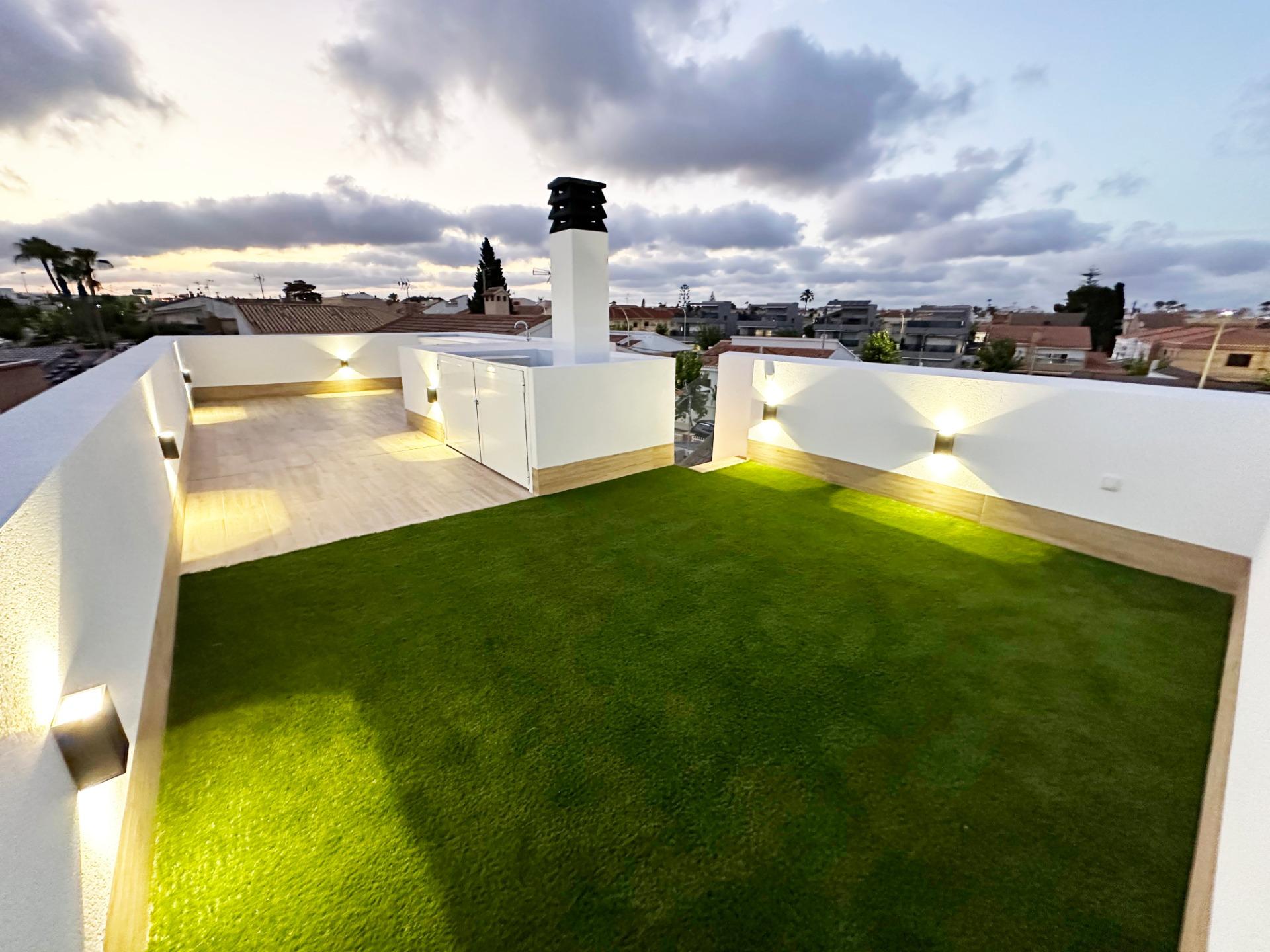 Rooftop terrace with artificial grass, white walls, and warm wall lights, silhouetted chimney against a cloudy evening sky.