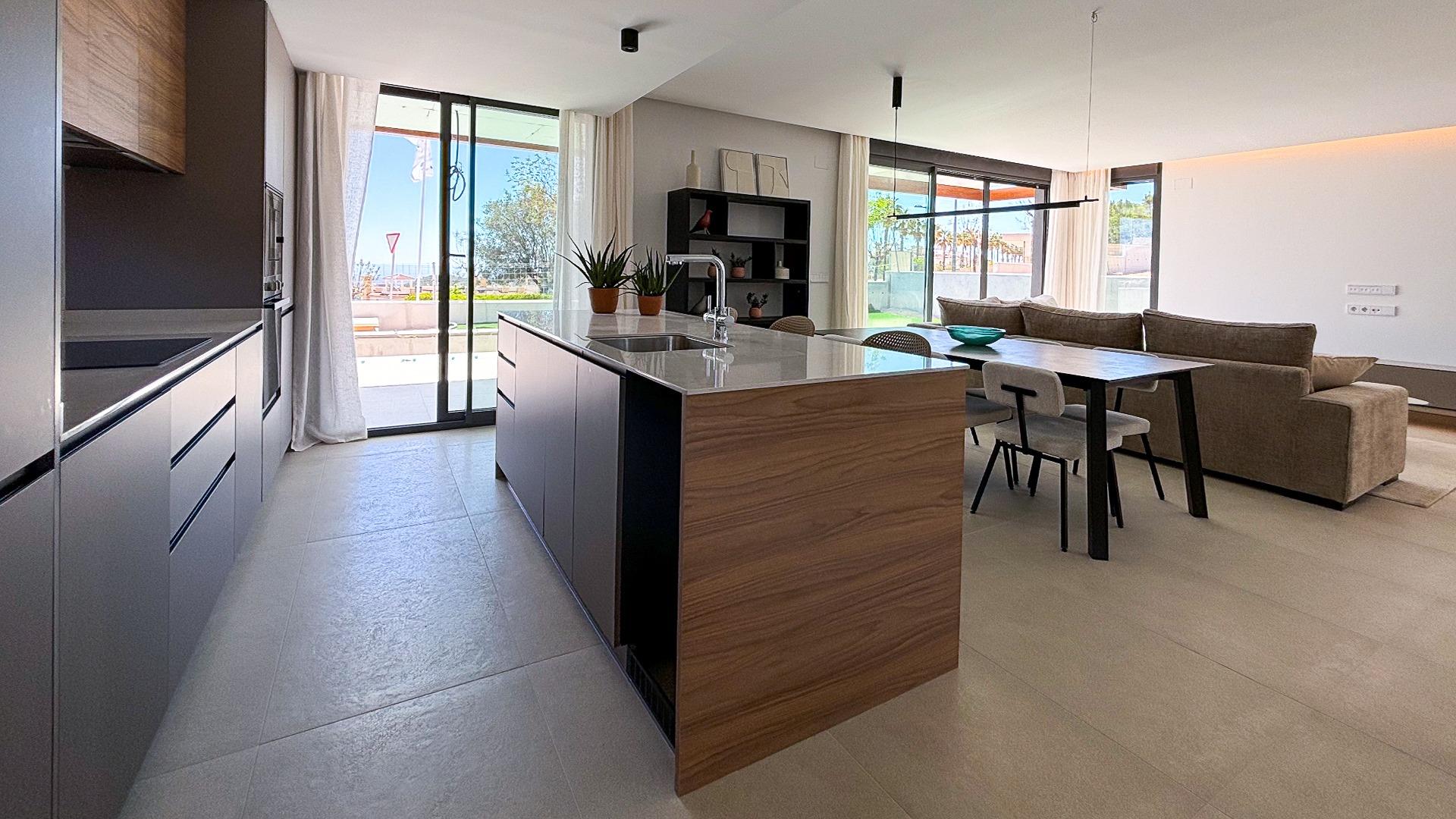 Open-plan kitchen with a large island, wood cabinets, and a dining table beside a beige sectional sofa in a sunlit living area.
