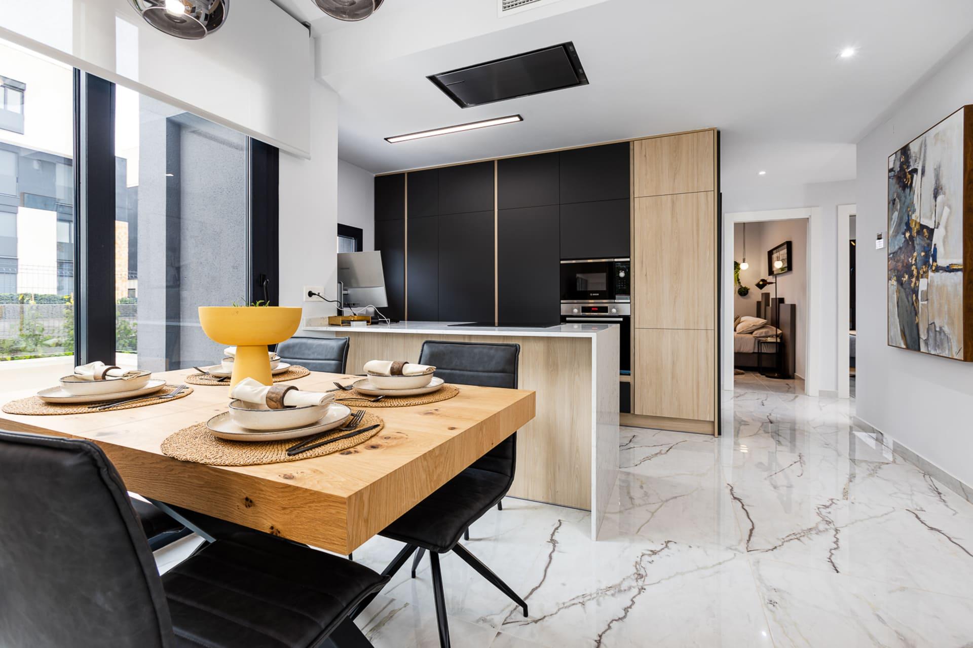 Open-concept kitchen and dining area with a wooden table set for six, black and wood cabinetry, and marble floors near large windows.