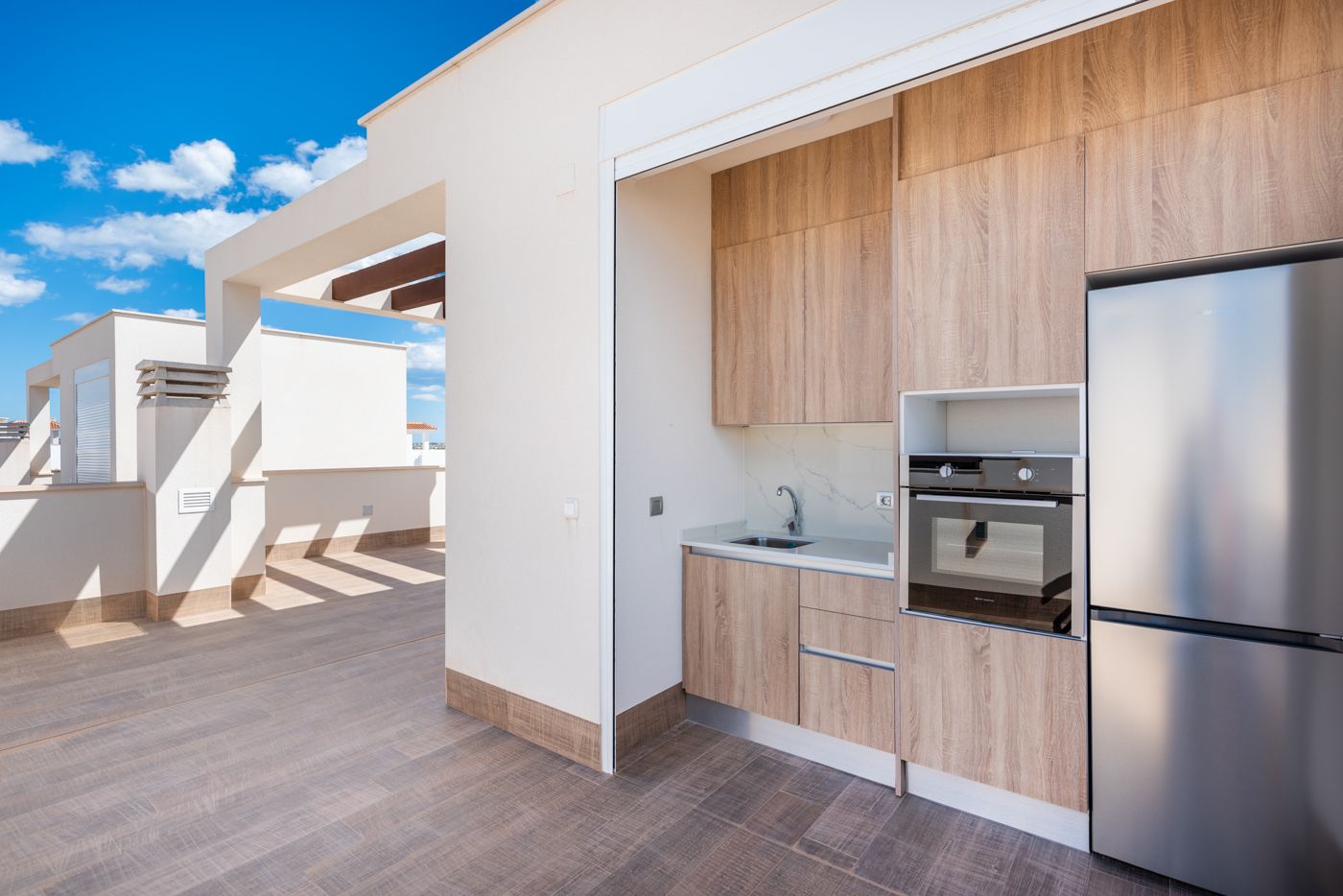 Modern outdoor kitchen with wooden cabinets, a stainless steel fridge and built-in oven on a sunlit terrace under a blue skyo.