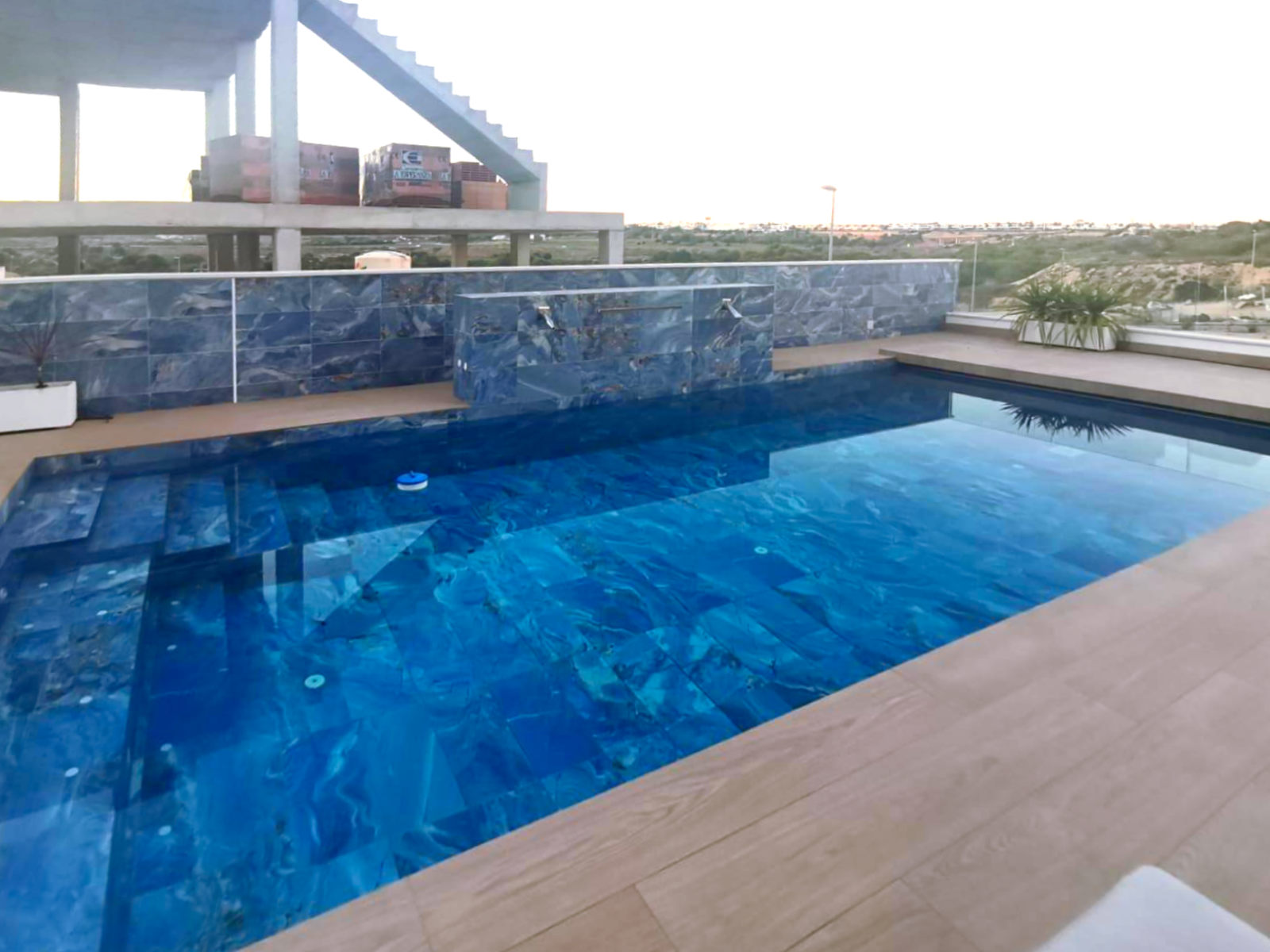 Rooftop swimming pool with blue tiled walls and wooden deck, cityscape visible in the distance.