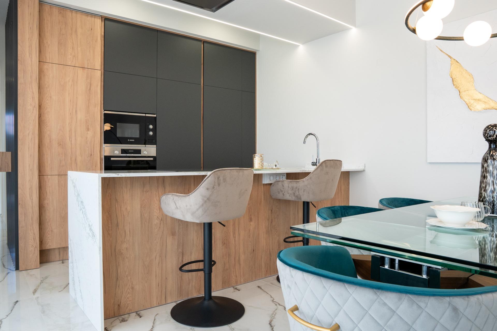 Modern kitchen with oak cabinetry, dark panel cupboards, built-in oven, marble island, and glass dining table with teal chairs.