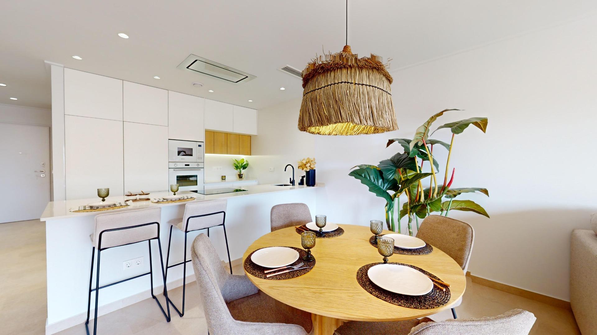 Open kitchen and dining area with a round wooden table set for four, woven pendant light, white cabinets, and a large plant in the corner.