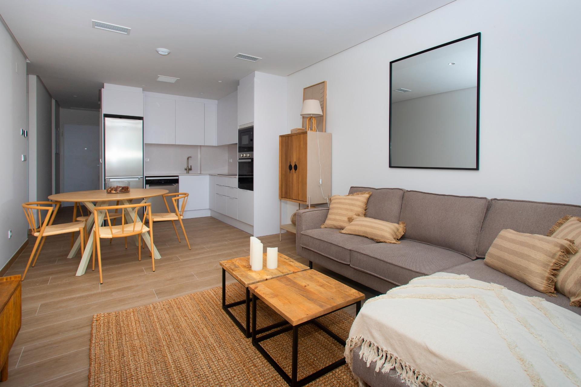 Open-plan living room and kitchen: gray sectional sofa, round wooden coffee table, woven rug, white cabinets and stainless appliances.