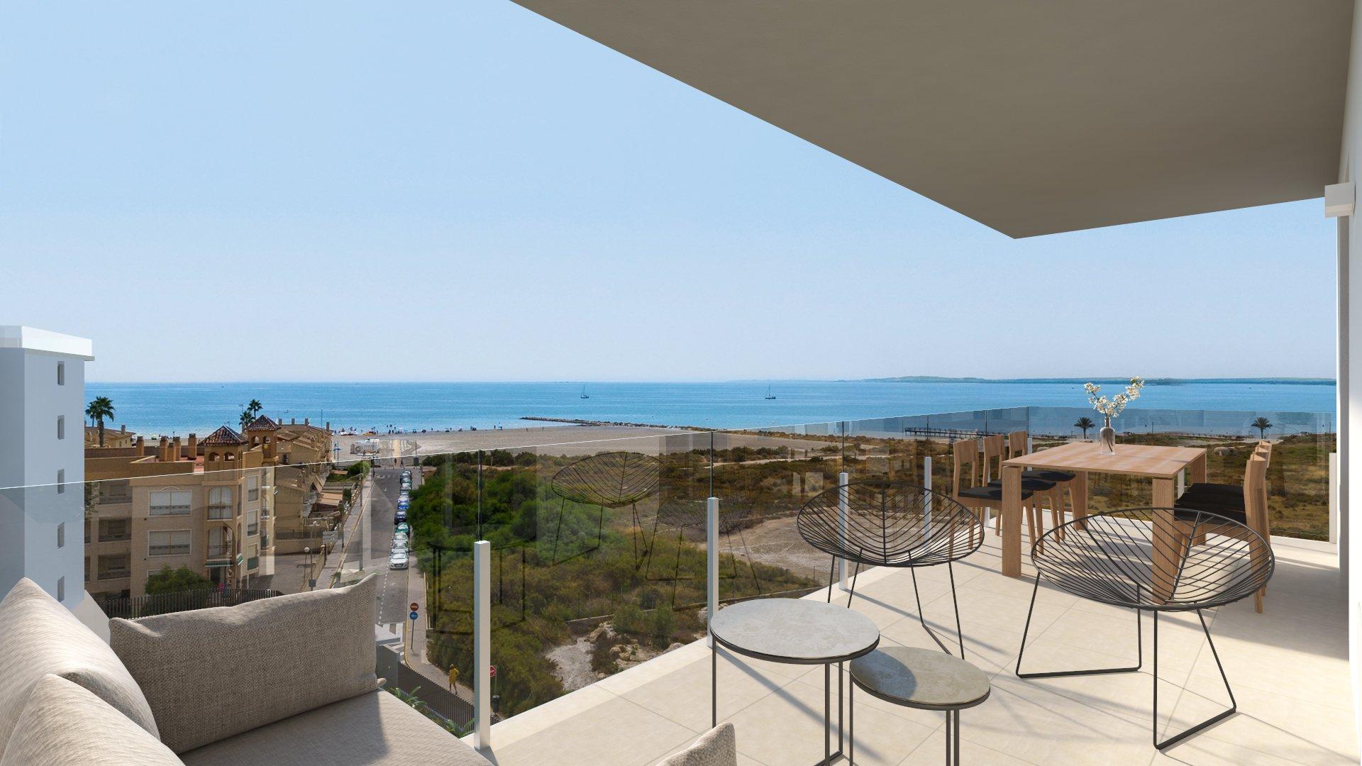 Outdoor balcony with glass railing, wicker chairs and a wooden table overlooking a sandy beach and blue ocean beyond.