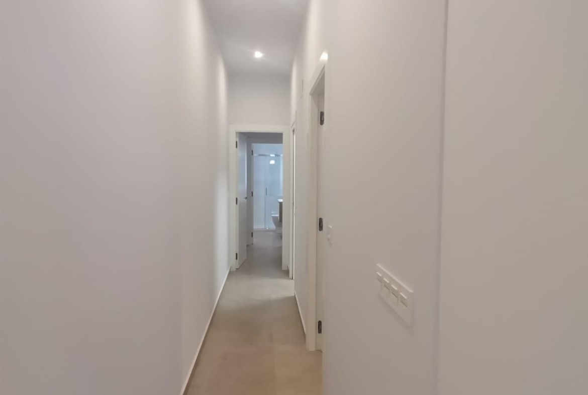 White, narrow hallway with doors on both sides and a bright room visible at the far end.