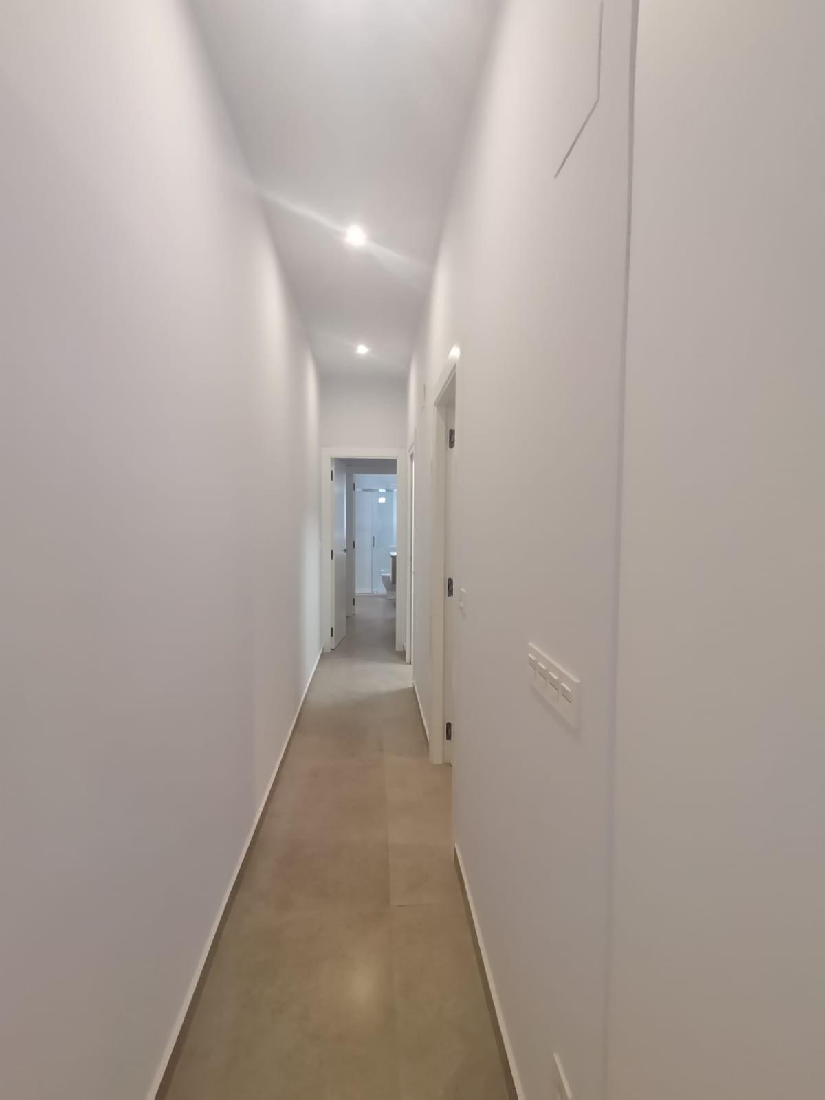 White, narrow hallway with doors on both sides and a bright room visible at the far end.