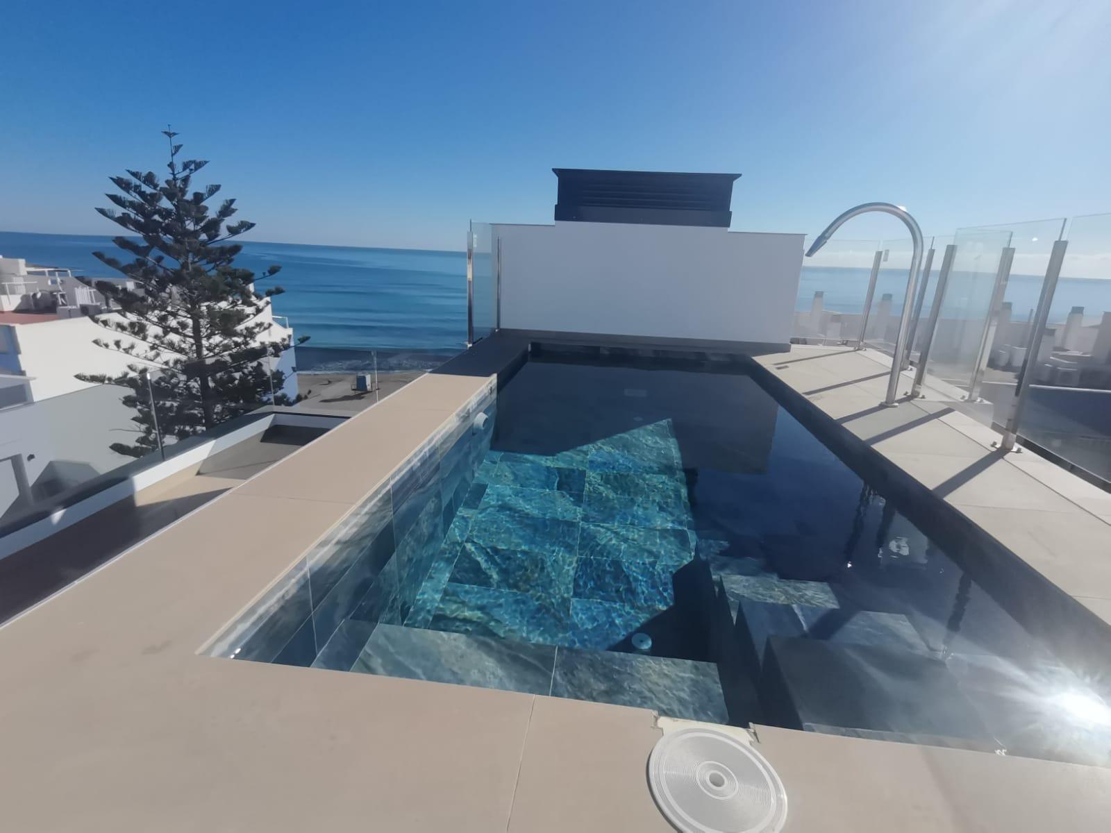 Rooftop infinity pool with clear blue water, glass barrier, and ocean view under a bright blue sky.