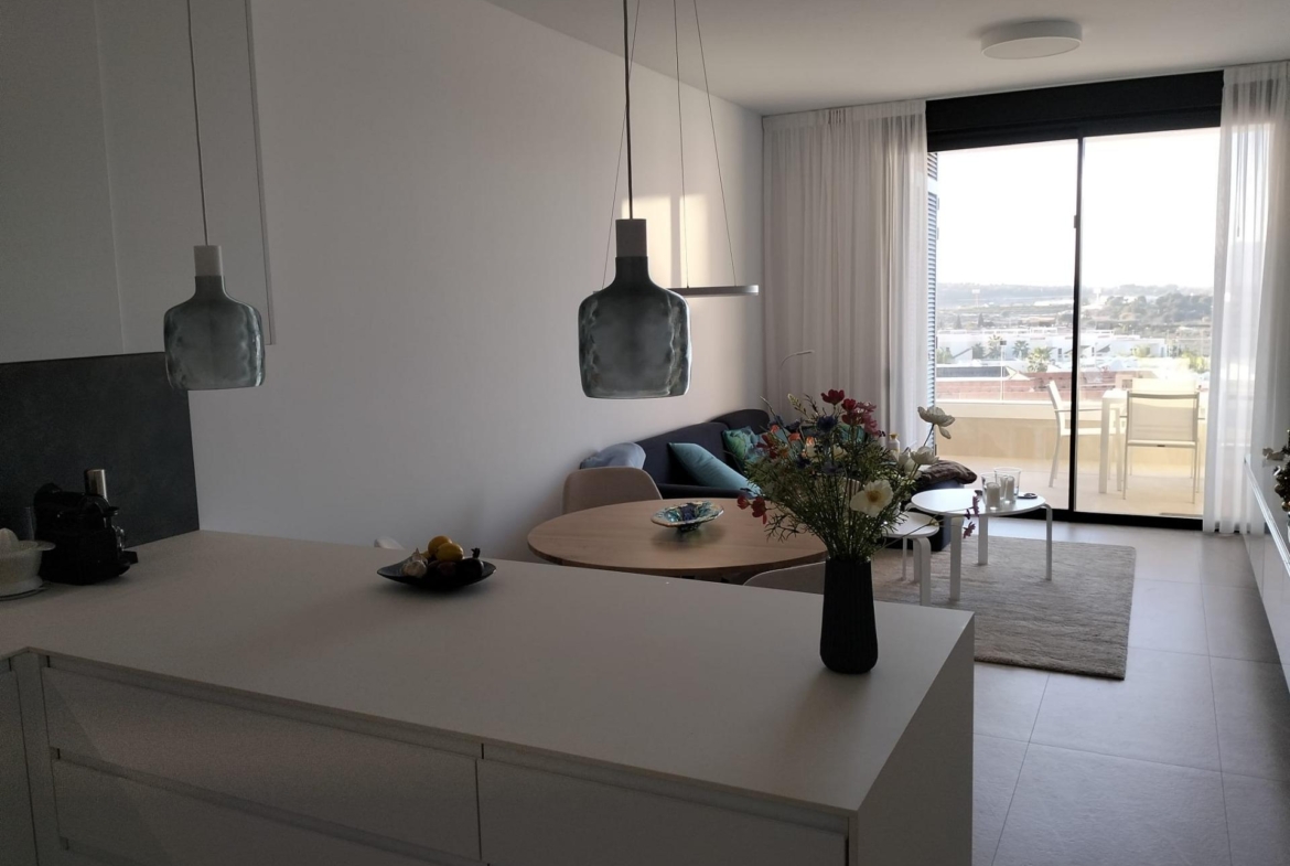 Bright open-plan kitchen and living area with white island, hanging pendant lights, and a vase of flowers on the island.