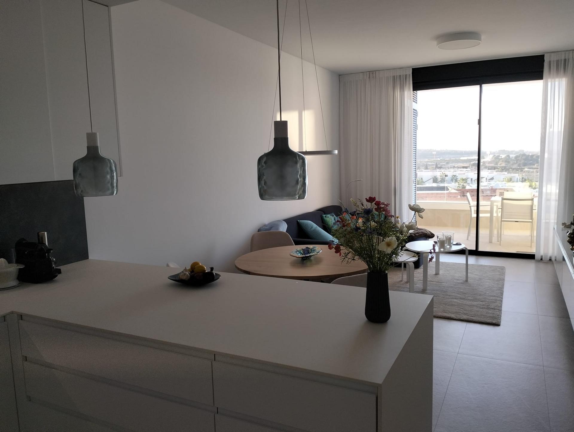 Bright open-plan kitchen and living area with white island, hanging pendant lights, and a vase of flowers on the island.