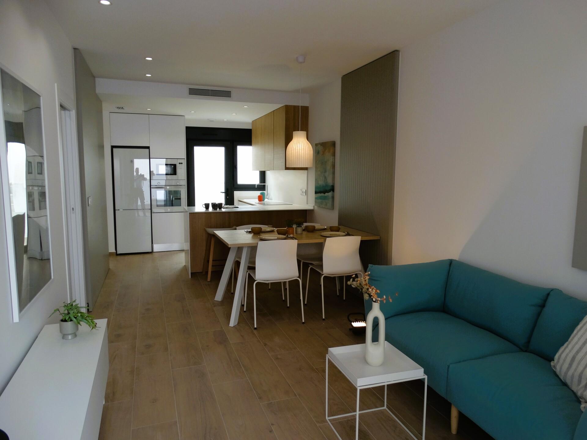 Open-plan kitchen and dining area with a wooden floor, white cabinetry, and a teal sofa nearby under a hanging pendant light.