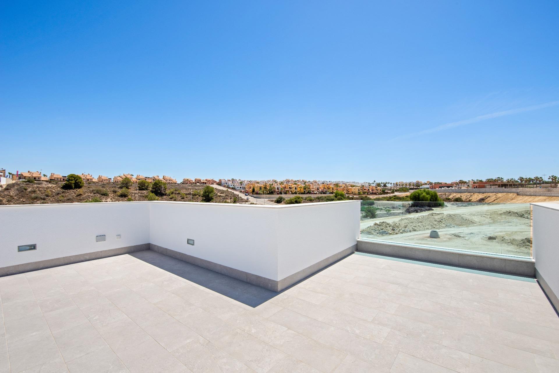Rooftop terrace with white walls and pale tile flooring, overlooking a hillside neighborhood under a clear blue sky.